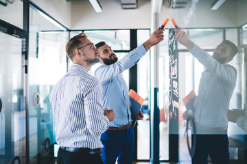 Confident male employee pointing on notes standing with colleague near glass creating infographic...