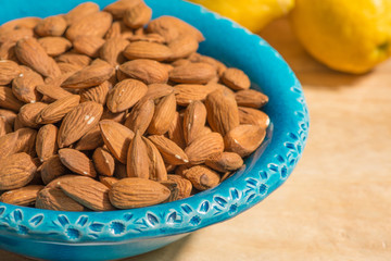 Closeup of shelled sicilian almonds in a blue bowl, lemons on background, selective focus.