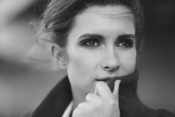 A beautiful lonely woman in a coat and scarf under a frozen bridge holds her collar with her hands. The concept of loneliness. Black and white art photo. Soft focus.