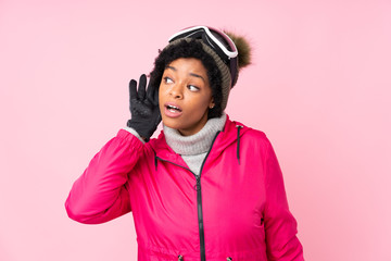 African american skier woman with snowboarding glasses over isolated pink background listening something