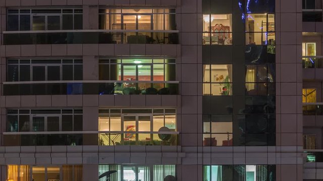 Night view of exterior apartment building timelapse. High rise skyscraper with blinking lights in windows with people moving inside. Pan down