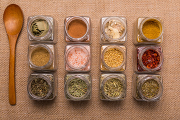 spices and aromatic herbs of different colors and textures in lined glass jars, on a brown background, with a wooden spoon