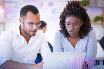 Diverse colleagues with laptop and virtual statistic graphics. Young man and woman using computer in office, looking at screen and talking. Corporate discussion concept