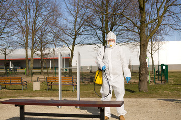 Osoba w stroju ochronnym spryskuje ławki w parku środkiem dezynfekującym by uniknąć  - obrazy, fototapety, plakaty