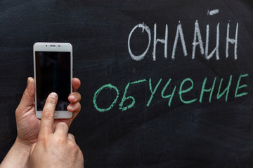 online education in Russian text in white and green chalk on the chalk Board. Hand with a mobile phone. The concept of online education