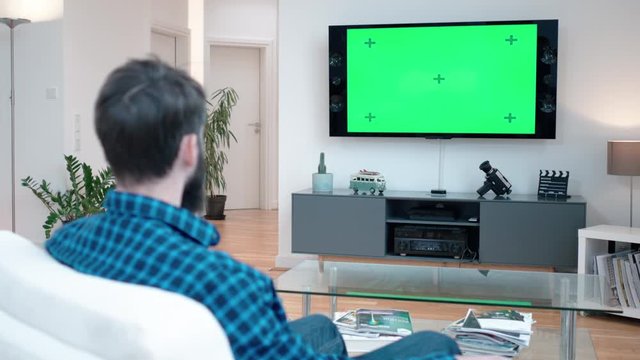 Man with a beard watching TV on a couch relaxing and switchs on TV with green screen shot in 4k