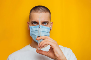 Portrait of a young guy in a respirator on a yellow background. Holding an ampoule with a coronavirus vaccine. Cold medicine. The concept of coronavirus.