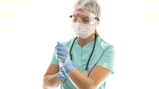 Female doctor in a medical mask and safety glasses puts on disposable gloves. Individual protection of medical workers.