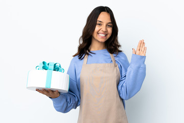 Pastry chef holding a big cake over isolated white background saluting with hand with happy expression