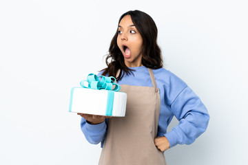 Pastry chef holding a big cake over isolated white background doing surprise gesture while looking to the side