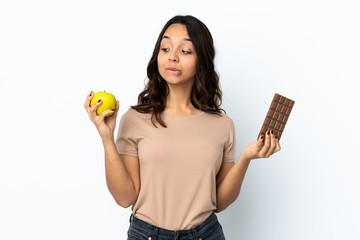 Young woman over isolated white background having doubts while taking a chocolate tablet in one...