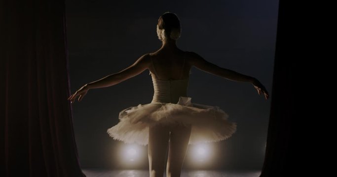 Theater curtains opens and Caucasian prima ballerina in tutu entering the stage and doing bowing pas for audience. Back view on female ballet dancer coming out and bow in darkness. Rear.
