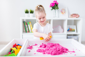Little blonde girl playing with pink kinetic sand on a white table in a light room. Sensory...