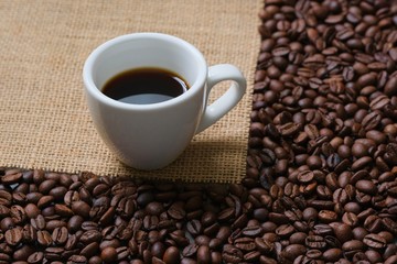 A white Cup of coffee on a background of coffee beans and coarse burlap. Coffee composition.