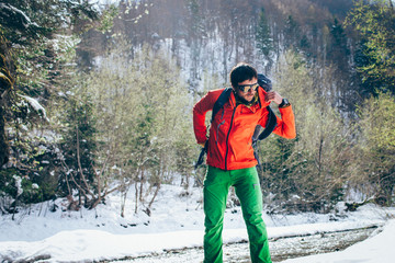 Fototapeta na wymiar Young male tourist takes off his backpack near a river in the mountains..Beautiful winter landscape with snow covered banks and trees on background. Climbing, hiking, trekking, active life concept.