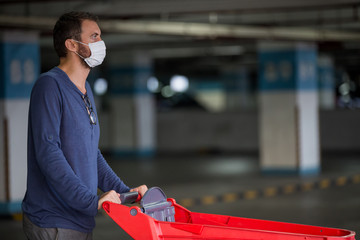 Young man wears medical face mask against corona virus covid-19 while pushing shopping cart outside of a grocery store in empty parking garage. Food supplies shortage. Panic buying and hoarding.