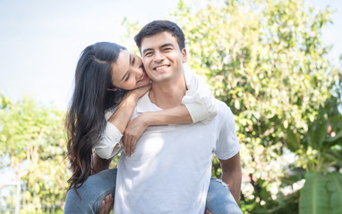 Naklejka premium Portrait of cute romantic caucasian guy in checkered shirt, dreamy asian lady rides him on rear. Leisure, chill happiness, lawn stroll, relax, romance lifestyle, well dressed partners posing