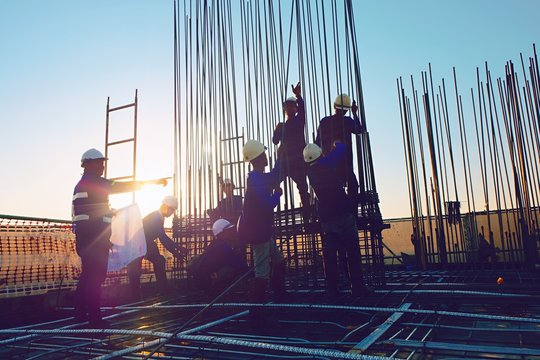 The construction of a rooftop high rise building, workers are tied with steel bars for concrete pouring, surveying the site and cranes are lifting or moving metal at the construction site. Pastel tone