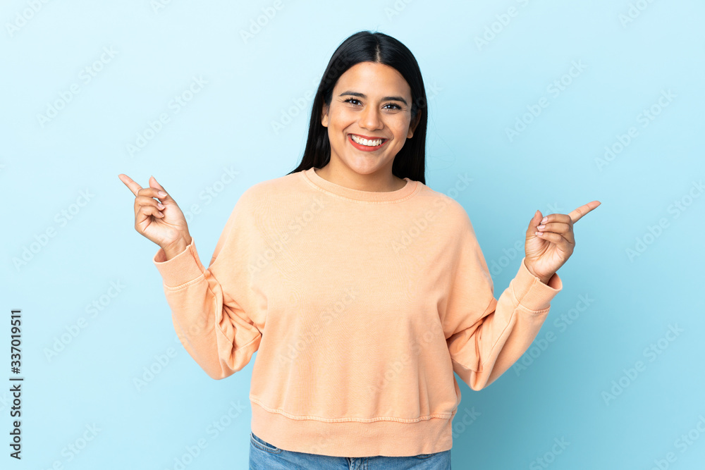 Wall mural Young latin woman woman isolated on blue background pointing finger to the laterals and happy