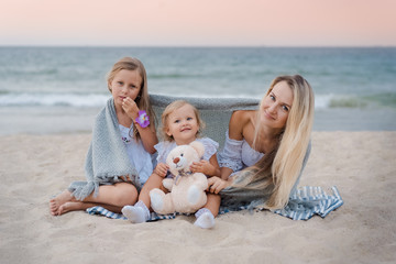 Mom with daughters blondes in white dresses laugh, hug and sit near the blue sea on the beach at sunset and hide behind a gray knitted blanket. Little girl holding a teddy bear.