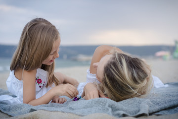 Mom with daughters blondes in white dresses laugh, hug and lying on a gray knitted blanket near the blue sea on the beach at sunset.