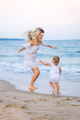 A blonde mother in a white dress plays with her daughter in the waves near the blue sea and laughs. Blurred.