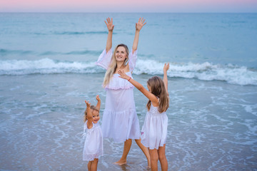 Mom with daughters blondes in white dresses laugh, hug and do exercises near the blue sea on the beach at sunset.