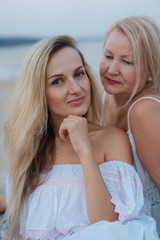 Blondes elderly mother and daughter in white dresses laugh, hug and sit near the blue sea on the beach at sunset.