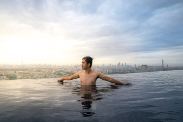Fototapeta premium Asian young man relaxing in the sky swimming pool watching cityscape 