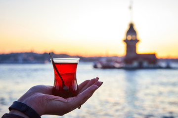 A cup (armud) of traditional Turkish tea in hand on the background of sunset over Bosphorus with famous Maiden's Tower (Kiz Kulesi ) also known as Leander's tower, symbol of Istanbul, Turkey