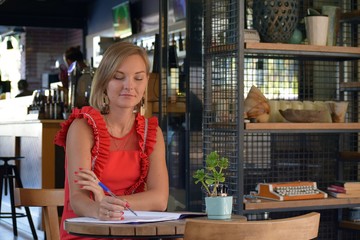 A sitting blonde girl in a red dress in a cafe writes.