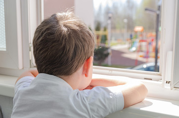 cute sad boy of four years old is sitting on the balcony and looking out the window at the...