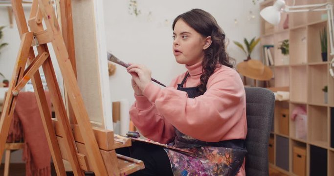 Teen wearing artists apron painting and putting brush from one hand to other at her room. Girl with down syndrome holding palette on knees while creating picture in front of molbert.
