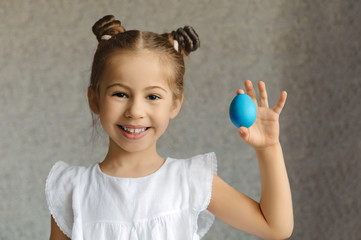 Happy girl with gathered dark hair in a white blouse on a gray background smiles and holds a blue Easter egg in his left hand