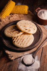 Arepas, South American food made with corn flour. Three arepas on a wooden plate and next to it a spoon and bowl full of cornmeal on a wooden background