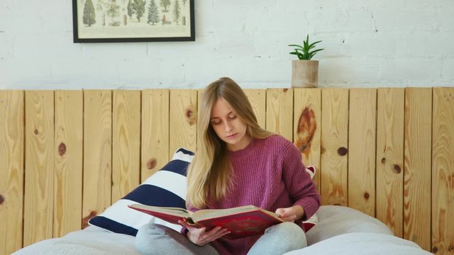 Young beautiful blonde woman in bed during sick leave or quarantine sits in bed. Wear comfortable and cosy outfit reads book. Studying remotely or relax at home during covid social distancing