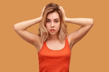 Beautiful blonde handsome girl tries to ignore annoying noise, plugs ears, has displeased expression. Young woman with shoulder length blonde hair wears t-shirt isolated on orange background in Studio