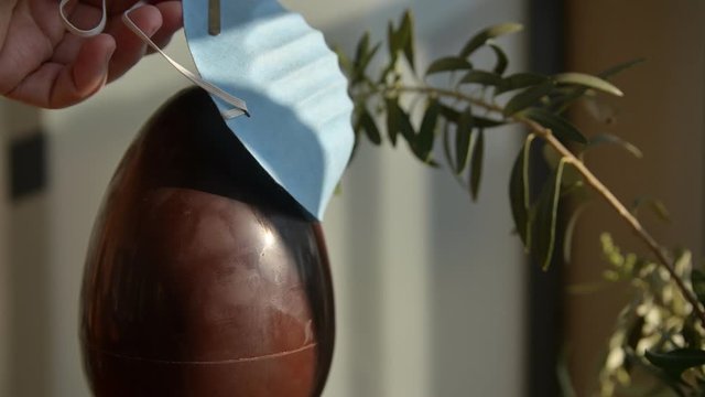Conceptual close-up image of Easter at the time of the coronavirus pandemic. A chocolate egg with an olive branch next to it. Caucasian man hands put a blue protective mask on the egg.