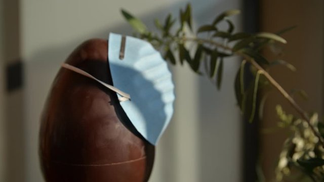 Conceptual close-up image of Easter at the time of the coronavirus pandemic. A chocolate egg with an olive branch next to it. Caucasian man hands put a blue protective mask on the egg.