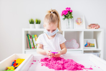 Little blonde girl playing with pink kinetic sand at home, wearing medical pritective face mask to protect herself from the virus