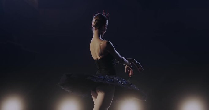 Back view on pretty young ballerina doing dancing pas in blue spotlight on stage during performance. Rear of classical ballet dancer in black tutu training and working hard.