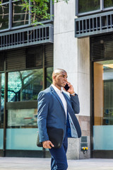 Fototapeta premium Young African American Business Man traveling, working in New York City, wearing blue blazer, white shirt, blue pants, carrying black briefcase, walking outside office building, talking on cell phone.