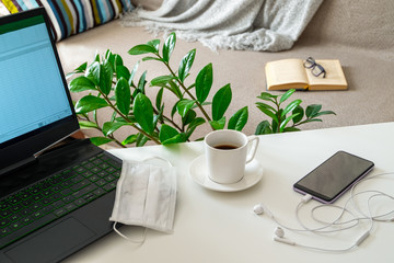 Workplace, home office with laptop, medical mask, gadgets and coffee. In the blurve background is a cozy home space. Remote work and education concept. Horizontal orientation. Selective focus.