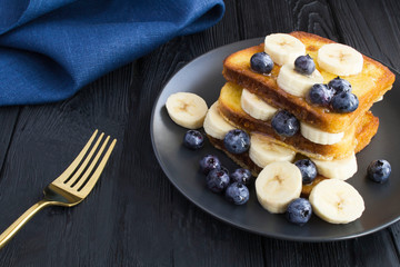 French toasts with blueberries, banana  and honey  in the black  plate on the dark wooden background. Closeup.