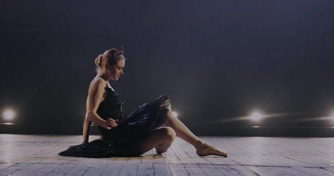 Caucasian young female ballet dancer in crown and black tutu sitting on stage and falling when trying to get up. Pretty ballerina starting to dance classical pas in studio with lights at rehearsal.