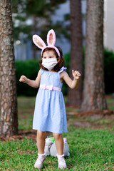 Little toddler girl with bunny ears and surgical face mask hunting for Easter eggs during coronavirus quarantine - 337056523