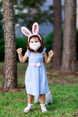 Little toddler girl with bunny ears and surgical face mask hunting for Easter eggs during coronavirus quarantine - 337056562