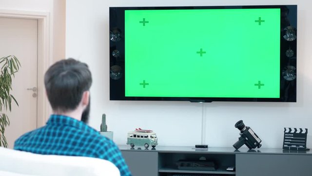 Man with a beard watching TV on a green screen while sitting on a couch in living room shot in 4k