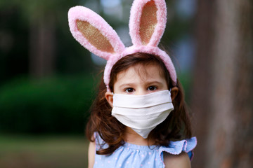 Little toddler girl with bunny ears and surgical face mask hunting for Easter eggs during coronavirus quarantine - 337056922