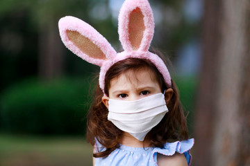 Little toddler girl with bunny ears and surgical face mask hunting for Easter eggs during coronavirus quarantine - 337056965
